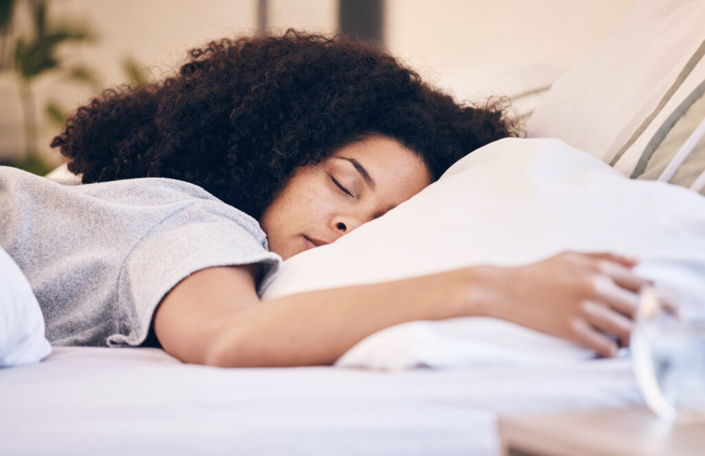 A peaceful woman with curly hair sleeping soundly on a comfortable bed, surrounded by soft pillows. The morning light gently illuminates the room, creating a calm and restful atmosphere.