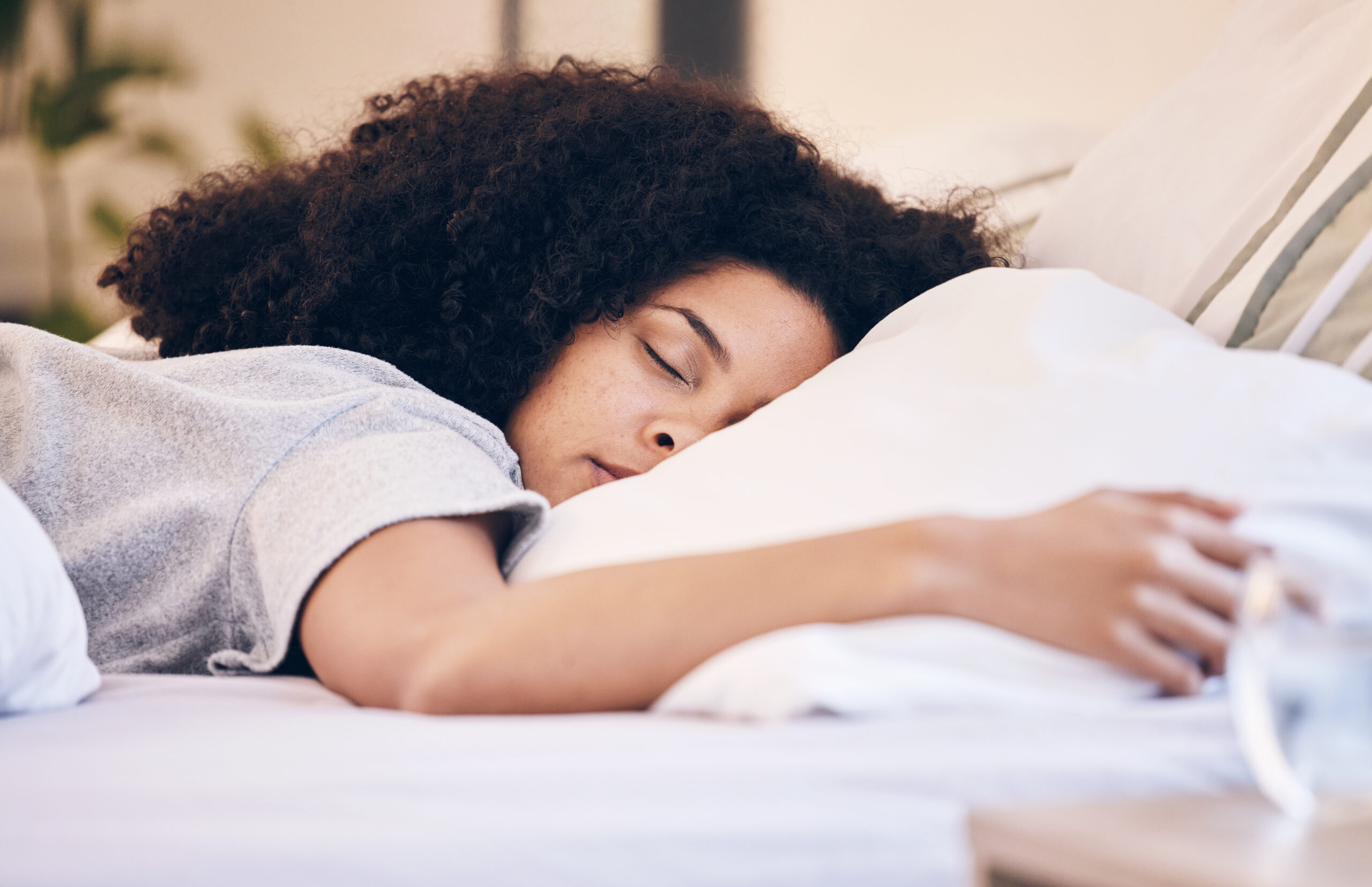 A peaceful woman with curly hair sleeping soundly on a comfortable bed, surrounded by soft pillows. The morning light gently illuminates the room, creating a calm and restful atmosphere.