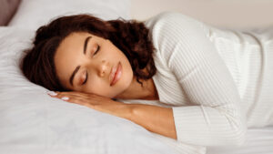 A peaceful young woman with curly hair sleeping soundly on a comfortable bed, resting her head on her hands. Her serene expression reflects a deep, restful sleep, surrounded by soft bedding.