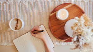 Top view of a woman writing in a lined journal beside a cup of coffee and a lit candle on a neutral textured surface.