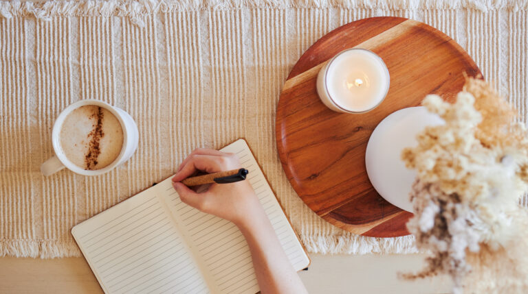 Top view of a woman writing in a lined journal beside a cup of coffee and a lit candle on a neutral textured surface.