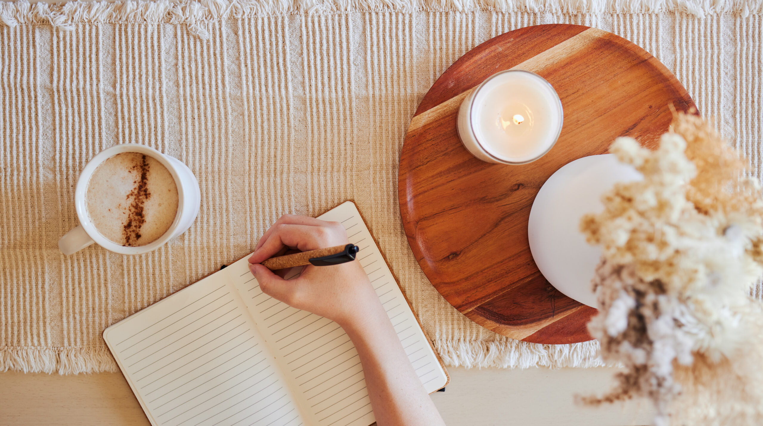 Top view of a woman writing in a lined journal beside a cup of coffee and a lit candle on a neutral textured surface.