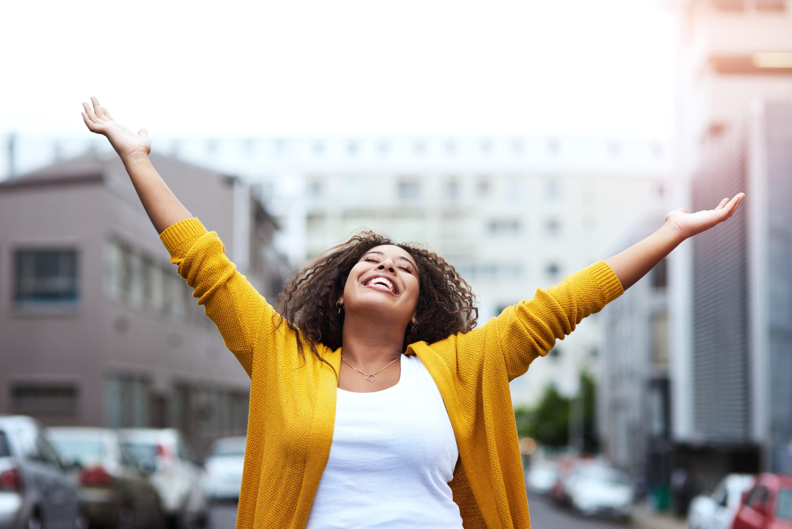 A woman with curly hair wearing a yellow cardigan joyfully spreading her arms outside, looking free and confident in an urban setting.