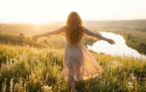 A woman with long, curly hair, seen from behind, stands in a sunlit field of wildflowers at sunset with her arms outstretched, overlooking a river valley.