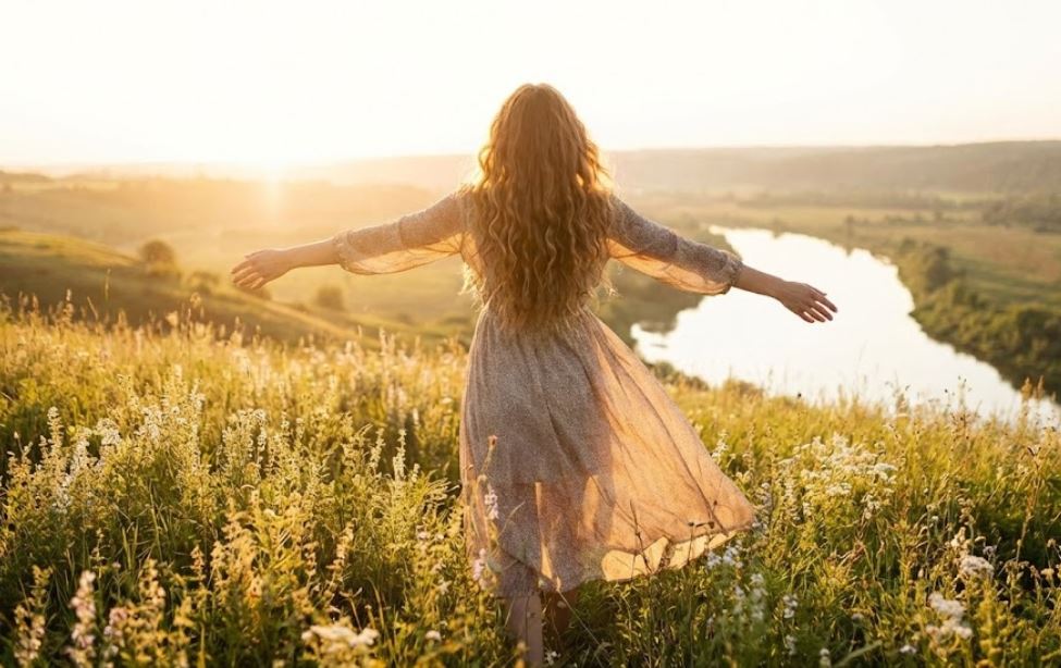 A woman with long, curly hair, seen from behind, stands in a sunlit field of wildflowers at sunset with her arms outstretched, overlooking a river valley.