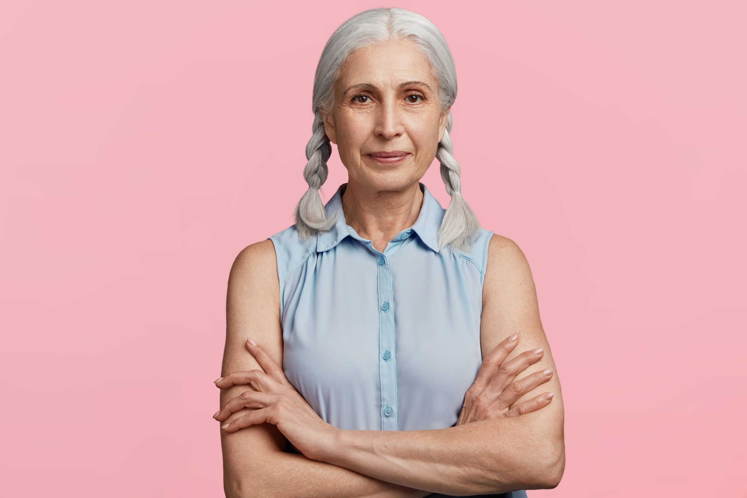 An older woman with gray hair and braids smiling confidently, standing against a pink background.