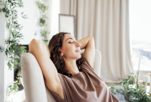 Smiling woman with curly hair sitting on a green couch, with her hand resting on her cheek, eyes closed.